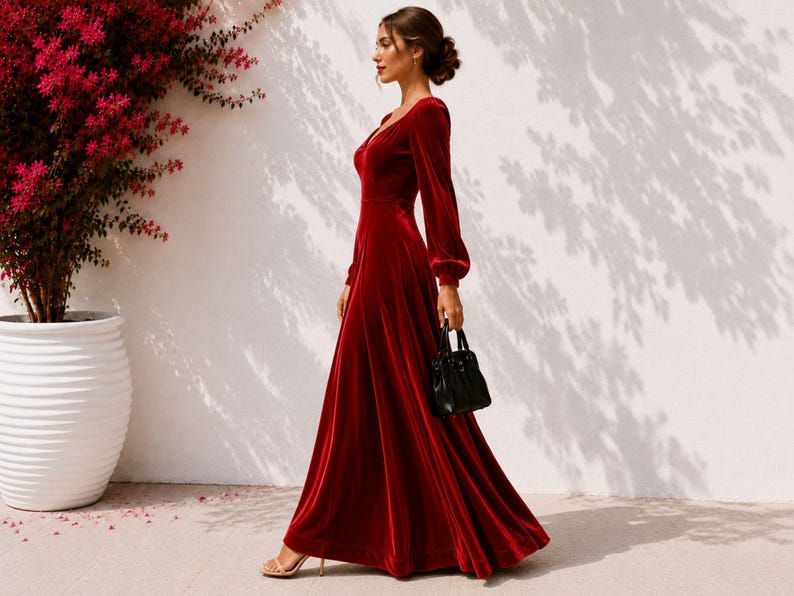 May include: A floor-length deep red velvet dress with long sleeves, a fitted bodice, and a flowing skirt. The dress features a V-neckline and cuffed sleeves. A small black handbag is held at the side, and beige strappy high-heeled sandals are worn.