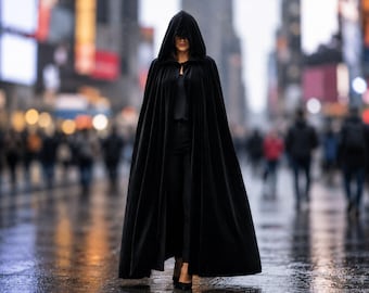 May include: A floor-length black velvet cloak featuring a deep hood that covers the head. The garment is fastened at the neck with a simple clasp and drapes down to the floor, creating a full silhouette over a black shirt and black trousers.