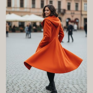 May include: An orange hooded coat with a flowing skirt is worn by a person in a city square. The coat is a vibrant shade of orange, and the person is wearing black pants and black boots. The coat's skirt is in motion.