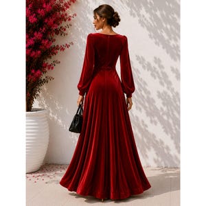 May include: A floor-length, deep red velvet dress featuring long, full sleeves with fitted cuffs, a defined waistline, and a flowing, pleated skirt. The garment has a simple, solid-colored design with a smooth texture and a back zipper closure.