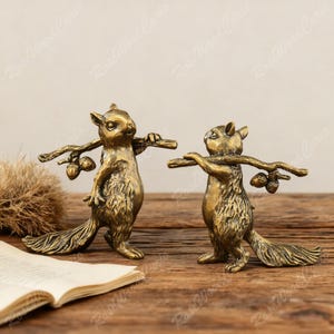 May include: Two bronze squirrel ornaments, each carrying a branch with acorns. The squirrels have detailed fur and long, bushy tails. The ornaments are displayed on a wooden surface, with a book and dried plant in the background.