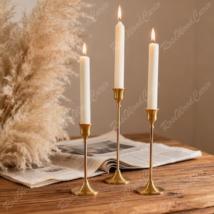 May include: Three lit white candles in gold-colored metal candlesticks. The candlesticks are of varying heights and are placed on a wooden surface with a newspaper and dried ornamental grass in the background. The candles are lit with a soft glow.