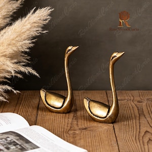 Könnte beinhalten: Zwei goldfarbene Schwanfiguren mit langen Hälsen und geschwungenen Körpern. Die Schwäne werden auf einer Holzoberfläche präsentiert, mit einem Buch und getrockneten Pflanzen im Hintergrund. Die Figuren haben eine Vintage-Ästhetik.