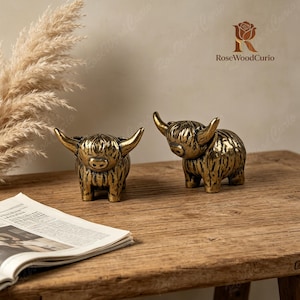 May include: Two metallic gold-toned highland cow figurines with textured fur details and prominent horns. The figures are standing side by side, each featuring a rounded body, short legs, and a distinct snout. The brand name RoseWoodCurio is visible.