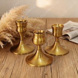 May include: Three brass candle holders with a warm, golden finish. Each holder has a different design, with a flared base and a cup-shaped top to hold candles. The candle holders are arranged on a wooden surface.