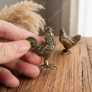 May include: A detailed bronze-colored rooster figurine held by a hand, with a matching hen figurine in the background. The rooster is in a crowing pose, showcasing intricate feather details. The figurines are on a wooden surface.