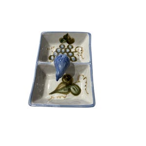 Op de afbeelding: Een keramische serveerschaal met twee niveaus en een blauwe vijgvormige handgreep. De schaal is versierd met geschilderde druiven en bladeren in groen- en bruintinten, met een blauwe rand. De schaal is vierkant met afgeronde hoeken.