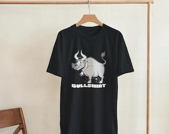 May include: Black t-shirt featuring a cartoon bull with a bell around its neck. The bull is gray and white with a smiling expression. The text "BULLSHIRT" is printed in white below the bull.