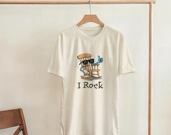May include: Cream-colored t-shirt featuring a cartoon rocking chair character wearing sunglasses and giving a thumbs-up. The text "I Rock" is printed below the graphic. The shirt is hanging on a wooden clothing rack.