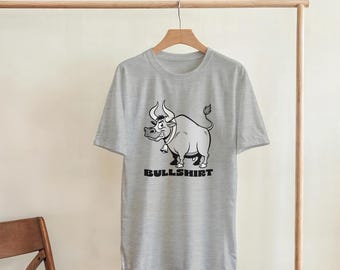 May include: A light gray t-shirt featuring a cartoon bull with a bell around its neck. The bull is in black and white, with the word "BULLSHIRT" printed below it in a bold, sans-serif font.