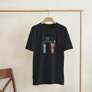 Puede incluir: Camiseta negra con un gráfico humorístico de temática científica. El diseño presenta dos tubos de ensayo de dibujos animados, uno con líquido azul y otro con líquido rojo, y el texto "I think you're overreacting" en una burbuja.