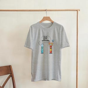 Puede incluir: Camiseta gris claro con un gráfico humorístico de dos tubos de ensayo. Uno es azul y tranquilo, el otro es rojo y explota. El texto de arriba dice "I think you're overreacting."