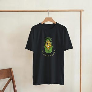 Op de afbeelding: Zwart T-shirt met een cartoon maïs ontwerp. Het ontwerp bevat een knipogende maïskarakter, groene bladeren en de tekst "A Little Corny". Het T-shirt hangt aan een houten kleerhanger.