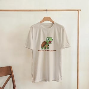 Op de afbeelding: Lichtbeige T-shirt met een cartoon schildpad ontwerp. De schildpad heeft een bruin schild en groene huid, met een verbaasde uitdrukking. De tekst "Shell Shocked" is eronder in bruin gedrukt.