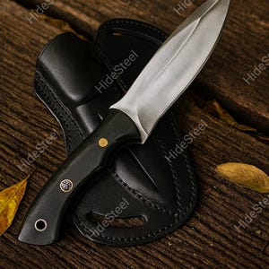 May include: A handcrafted hunting knife with a polished steel blade and a black handle, resting on a black leather sheath. The knife features a sharp point and is set against a rustic wooden background.