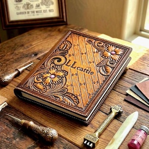 May include: A tan leather journal with intricate floral tooling and the text LL cattle embossed on the cover. The journal is surrounded by leatherworking tools including two metal awls, a metal stamp, a bone folder, and a small bottle of pink dye.