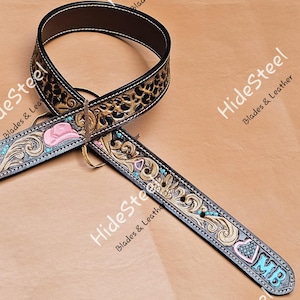 Könnte beinhalten: Ein brauner Gitarrengurt aus Leder mit Leopardenmuster und weißen Nähten. Der Gurt hat einen rosa Cowboyhut, türkisfarbene Akzente und die Initialen "MB" in Blau.
