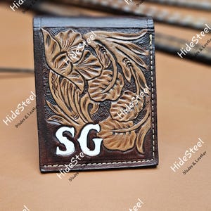 May include: A brown leather wallet with floral carving and the initials "SG" in white. The wallet is rectangular and features a detailed embossed design. The background is a light brown leather surface.