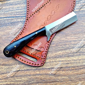 May include: A straight-edged knife with a black handle and a silver blade rests on a brown leather sheath. The sheath has decorative stitching and a cutout. The knife and sheath are on a wooden surface.