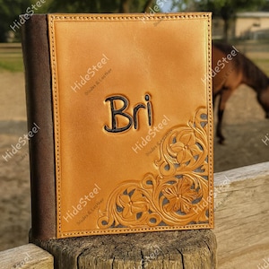 May include: A tan leather journal cover featuring a dark brown spine and a hand-tooled floral pattern in the bottom right corner. The name Bri is embossed in black lettering in the center of the front cover, surrounded by decorative stitching.