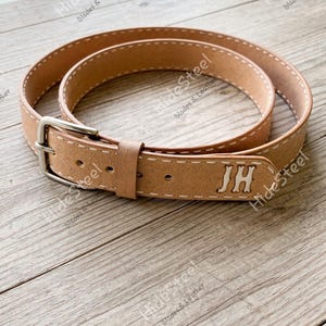May include: A tan leather belt with white stitching and a silver buckle. The belt is personalized with the initials "JH". The words "HideSteel Blades & Leather" are printed on the belt.