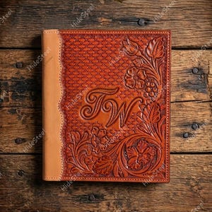 May include: A rectangular leather journal with a tan spine and a dark brown cover featuring intricate floral tooling, a cross-hatched background pattern, and the embossed letters TW in the center, all enclosed within a decorative stitched border.