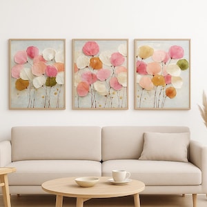 Peut inclure: Trois impressions d'art floral encadrées. Chaque impression présente des formes de fleurs abstraites et colorées en rose, pêche, blanc et vert. L'œuvre est accrochée au-dessus d'un canapé beige et d'une table basse ronde.