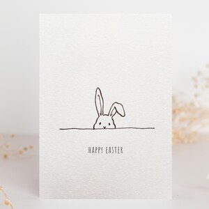 May include: A white Easter card featuring a minimalist line drawing of a rabbit's head and ears. The words "HAPPY EASTER" are printed in a simple font below the rabbit. The design is a simple black line drawing on a textured white background.