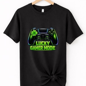 Pode incluir: Camiseta preta com um gráfico de controle de videogame verde neon e azul. O controle tem um trevo de quatro folhas e o texto "LUCKY GAMER MODE" em verde. A camiseta está pendurada em um cabide de madeira.
