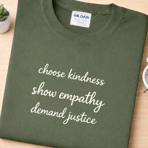 Puede incluir: Camiseta verde oliva con las palabras "choose kindness, show empathy, demand justice" en escritura blanca. La camiseta está doblada sobre una superficie de madera clara. Una pequeña planta en maceta blanca es visible en la esquina superior izquierda.