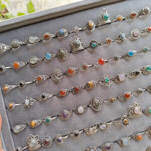 Hand holding a rectangular grey velvet jewelry display tray filled with multiple rows of small minimalist silver-toned gemstone rings. Each ring features delicate, dainty designs with tiny oval, round, square, and teardrop stones in colors like turquoise, carnelian, amethyst, moonstone, and green onyx. The rings have subtle vintage-inspired details and thin bands, arranged neatly in horizontal lines. The photo is taken outdoors in natural daylight with soft greenery.
