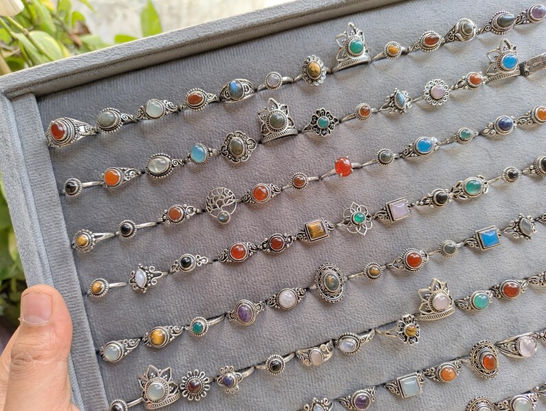 Hand holding a rectangular grey velvet jewelry display tray filled with multiple rows of small minimalist silver-toned gemstone rings. Each ring features delicate, dainty designs with tiny oval, round, square, and teardrop stones in colors like turquoise, carnelian, amethyst, moonstone, and green onyx. The rings have subtle vintage-inspired details and thin bands, arranged neatly in horizontal lines. The photo is taken outdoors in natural daylight with soft greenery.