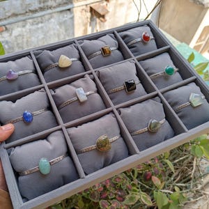 May include: A display of silver bracelets, each featuring a different gemstone. The gemstones vary in color, including purple, yellow, brown, red, green, black, blue, and clear. The bracelets are arranged in a gray display case.