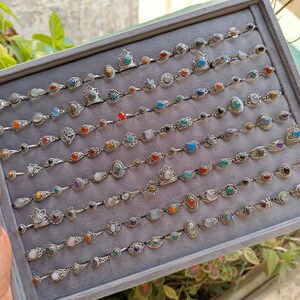 Hand holding a rectangular grey velvet jewelry display tray filled with multiple rows of small minimalist silver-toned gemstone rings. Each ring features delicate, dainty designs with tiny oval, round, square, and teardrop stones in colors like turquoise, carnelian, amethyst, moonstone, and green onyx. The rings have subtle vintage-inspired details and thin bands, arranged neatly in horizontal lines. The photo is taken outdoors in natural daylight with soft greenery.