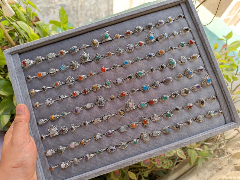 Hand holding a rectangular grey velvet jewelry display tray filled with multiple rows of small minimalist silver-toned gemstone rings. Each ring features delicate, dainty designs with tiny oval, round, square, and teardrop stones in colors like turquoise, carnelian, amethyst, moonstone, and green onyx. The rings have subtle vintage-inspired details and thin bands, arranged neatly in horizontal lines. The photo is taken outdoors in natural daylight with soft greenery.