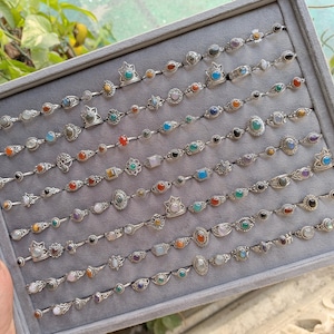Hand holding a rectangular grey velvet jewelry display tray filled with multiple rows of small minimalist silver-toned gemstone rings. Each ring features delicate, dainty designs with tiny oval, round, square, and teardrop stones in colors like turquoise, carnelian, amethyst, moonstone, and green onyx. The rings have subtle vintage-inspired details and thin bands, arranged neatly in horizontal lines. The photo is taken outdoors in natural daylight with soft greenery.