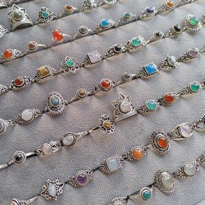 Hand holding a rectangular grey velvet jewelry display tray filled with multiple rows of small minimalist silver-toned gemstone rings. Each ring features delicate, dainty designs with tiny oval, round, square, and teardrop stones in colors like turquoise, carnelian, amethyst, moonstone, and green onyx. The rings have subtle vintage-inspired details and thin bands, arranged neatly in horizontal lines. The photo is taken outdoors in natural daylight with soft greenery.