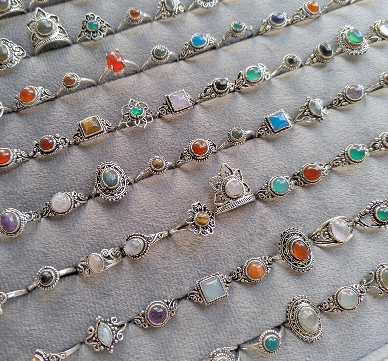 Hand holding a rectangular grey velvet jewelry display tray filled with multiple rows of small minimalist silver-toned gemstone rings. Each ring features delicate, dainty designs with tiny oval, round, square, and teardrop stones in colors like turquoise, carnelian, amethyst, moonstone, and green onyx. The rings have subtle vintage-inspired details and thin bands, arranged neatly in horizontal lines. The photo is taken outdoors in natural daylight with soft greenery.