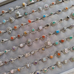 Hand holding a rectangular grey velvet jewelry display tray filled with multiple rows of small minimalist silver-toned gemstone rings. Each ring features delicate, dainty designs with tiny oval, round, square, and teardrop stones in colors like turquoise, carnelian, amethyst, moonstone, and green onyx. The rings have subtle vintage-inspired details and thin bands, arranged neatly in horizontal lines. The photo is taken outdoors in natural daylight with soft greenery.