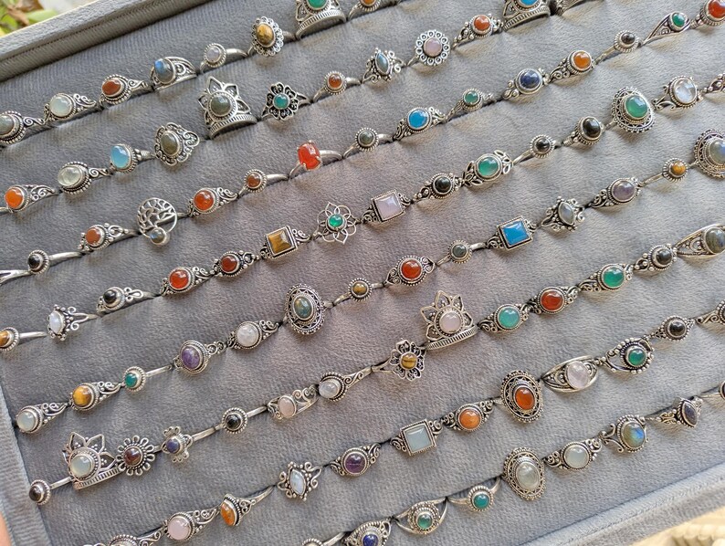Hand holding a rectangular grey velvet jewelry display tray filled with multiple rows of small minimalist silver-toned gemstone rings. Each ring features delicate, dainty designs with tiny oval, round, square, and teardrop stones in colors like turquoise, carnelian, amethyst, moonstone, and green onyx. The rings have subtle vintage-inspired details and thin bands, arranged neatly in horizontal lines. The photo is taken outdoors in natural daylight with soft greenery.