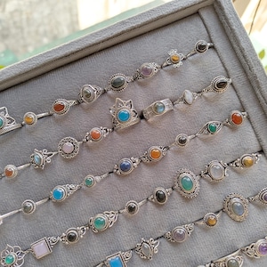 Hand holding a rectangular grey velvet jewelry display tray filled with multiple rows of small minimalist silver-toned gemstone rings. Each ring features delicate, dainty designs with tiny oval, round, square, and teardrop stones in colors like turquoise, carnelian, amethyst, moonstone, and green onyx. The rings have subtle vintage-inspired details and thin bands, arranged neatly in horizontal lines. The photo is taken outdoors in natural daylight with soft greenery.