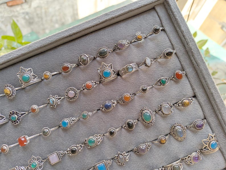 Hand holding a rectangular grey velvet jewelry display tray filled with multiple rows of small minimalist silver-toned gemstone rings. Each ring features delicate, dainty designs with tiny oval, round, square, and teardrop stones in colors like turquoise, carnelian, amethyst, moonstone, and green onyx. The rings have subtle vintage-inspired details and thin bands, arranged neatly in horizontal lines. The photo is taken outdoors in natural daylight with soft greenery.