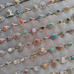 Hand holding a rectangular grey velvet jewelry display tray filled with multiple rows of small minimalist silver-toned gemstone rings. Each ring features delicate, dainty designs with tiny oval, round, square, and teardrop stones in colors like turquoise, carnelian, amethyst, moonstone, and green onyx. The rings have subtle vintage-inspired details and thin bands, arranged neatly in horizontal lines. The photo is taken outdoors in natural daylight with soft greenery.