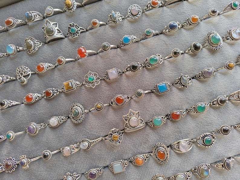 Hand holding a rectangular grey velvet jewelry display tray filled with multiple rows of small minimalist silver-toned gemstone rings. Each ring features delicate, dainty designs with tiny oval, round, square, and teardrop stones in colors like turquoise, carnelian, amethyst, moonstone, and green onyx. The rings have subtle vintage-inspired details and thin bands, arranged neatly in horizontal lines. The photo is taken outdoors in natural daylight with soft greenery.