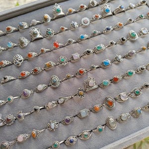 Hand holding a rectangular grey velvet jewelry display tray filled with multiple rows of small minimalist silver-toned gemstone rings. Each ring features delicate, dainty designs with tiny oval, round, square, and teardrop stones in colors like turquoise, carnelian, amethyst, moonstone, and green onyx. The rings have subtle vintage-inspired details and thin bands, arranged neatly in horizontal lines. The photo is taken outdoors in natural daylight with soft greenery.