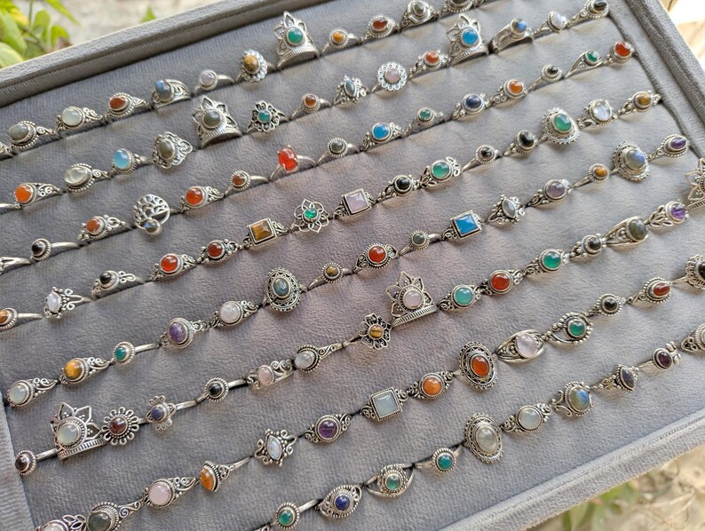 Hand holding a rectangular grey velvet jewelry display tray filled with multiple rows of small minimalist silver-toned gemstone rings. Each ring features delicate, dainty designs with tiny oval, round, square, and teardrop stones in colors like turquoise, carnelian, amethyst, moonstone, and green onyx. The rings have subtle vintage-inspired details and thin bands, arranged neatly in horizontal lines. The photo is taken outdoors in natural daylight with soft greenery.