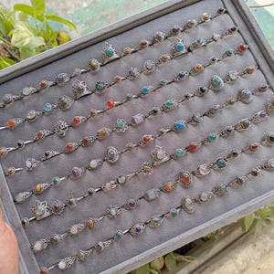Hand holding a rectangular grey velvet jewelry display tray filled with multiple rows of small minimalist silver-toned gemstone rings. Each ring features delicate, dainty designs with tiny oval, round, square, and teardrop stones in colors like turquoise, carnelian, amethyst, moonstone, and green onyx. The rings have subtle vintage-inspired details and thin bands, arranged neatly in horizontal lines. The photo is taken outdoors in natural daylight with soft greenery.
