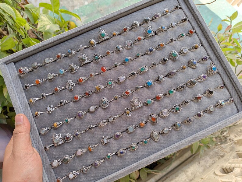 Hand holding a rectangular grey velvet jewelry display tray filled with multiple rows of small minimalist silver-toned gemstone rings. Each ring features delicate, dainty designs with tiny oval, round, square, and teardrop stones in colors like turquoise, carnelian, amethyst, moonstone, and green onyx. The rings have subtle vintage-inspired details and thin bands, arranged neatly in horizontal lines. The photo is taken outdoors in natural daylight with soft greenery.