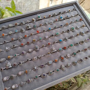 Hand holding a rectangular grey velvet jewelry display tray filled with multiple rows of small minimalist silver-toned gemstone rings. Each ring features delicate, dainty designs with tiny oval, round, square, and teardrop stones in colors like turquoise, carnelian, amethyst, moonstone, and green onyx. The rings have subtle vintage-inspired details and thin bands, arranged neatly in horizontal lines. The photo is taken outdoors in natural daylight with soft greenery.