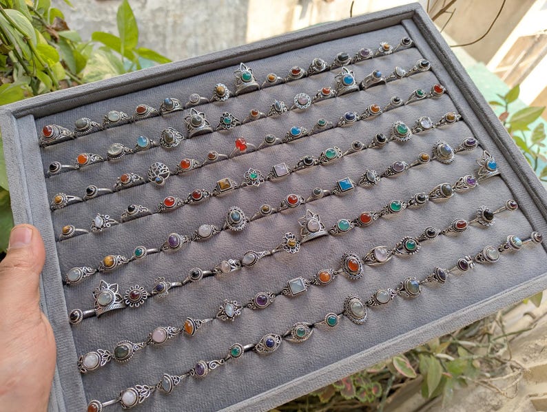 Hand holding a rectangular grey velvet jewelry display tray filled with multiple rows of small minimalist silver-toned gemstone rings. Each ring features delicate, dainty designs with tiny oval, round, square, and teardrop stones in colors like turquoise, carnelian, amethyst, moonstone, and green onyx. The rings have subtle vintage-inspired details and thin bands, arranged neatly in horizontal lines. The photo is taken outdoors in natural daylight with soft greenery.